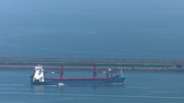 Large cargo ship sails on the blue ocean near a pier with trains a long