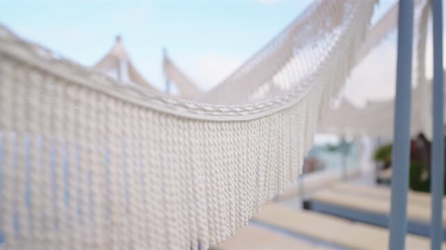 Close-up of elegant white cotton hammock with decorative fringe swaying gently at a beach resort in Santorini Greece. Soft focus background reveals thatched umbrellas and blue sky on a sunny day.