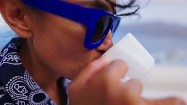 Close-up of a smiling Indian woman wearing stylish blue sunglasses sipping coffee from a white ceramic cup, dressed in a blue paisley patterned shirt against a soft blurred coastal background.