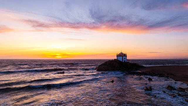 Aerial view of Capela do Senhor da Pedra on a rocky outcrop with waves crashing and a vibrant sunset sky Miramar, Porto, Portugal.