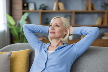 Obraz premium Mature blonde woman sitting comfortably on a modern sofa with hands behind her head, enjoying a moment of relaxation and tranquility in a cozy home environment