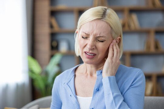 Mature woman suffering from an acute ear infection or earache, touching her painful ear and grimacing in agony while seeking relief and struggling with the discomfort