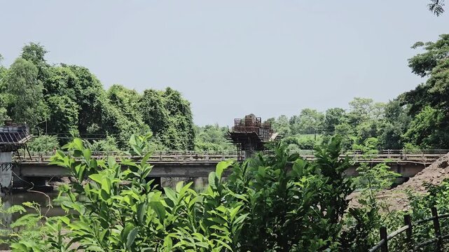 An excavator works on a dirt bank to construct a bridge pier over a river. The heavy machinery operates near a partially built structure and an older bridge. Green trees line the background under sky.