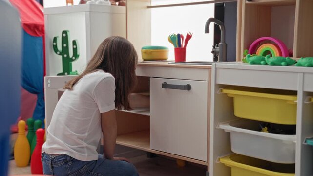 Little girl kneeling and reaching to a plastic bowl at a toy kitchen sink in a playroom building; playful curiosity.