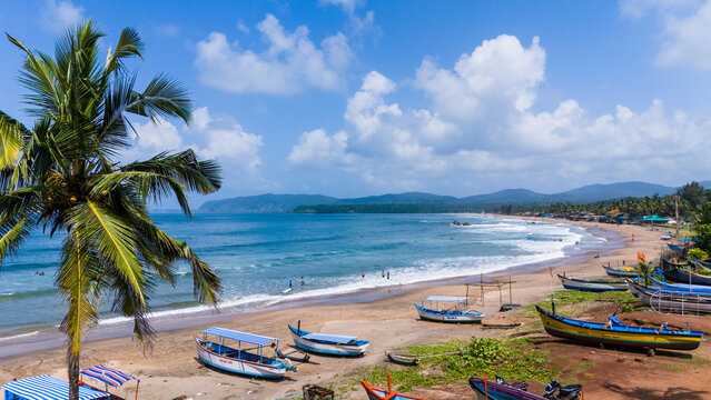 Aerial view of Agonda Beach with colorful fishing boats on the sandy shore, a palm tree, and turquoise waves under a cloudy sky Agonda, Goa, India.