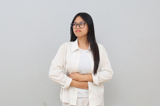 A young Asian woman is having stomach ache, bending over and holding her stomach, feeling uncomfortable due to menstrual cramps, stomach ache, diarrhea, standing isolated on white background.