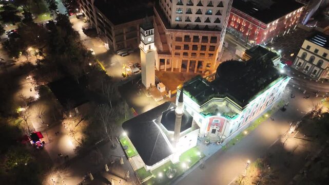 Aerial footage descending over Skanderbeg Square in Tirana, Albania, while panning the camera up past the illuminated Et'hem Bey Mosque, historic Clock Tower, and modern city buildings