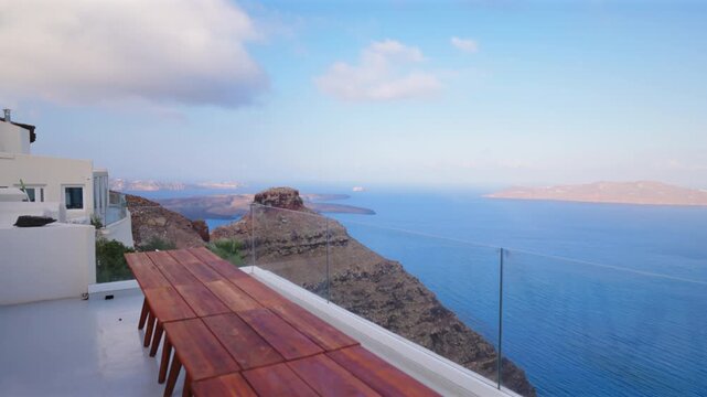 Stunning view from luxury villa terrace in Santorini Greece featuring wooden dining table with glass railing overlooking volcanic caldera and Aegean Sea with distant islands under cloudy sky.