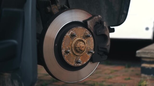 Front suspension turning with exposed brake disc rotor during car repair. Automotive brake system rotating, revealing wheel hub, caliper, rusty lug studs during maintenance. Vehicle maintenance