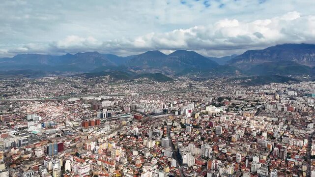 High-angle aerial perspective flying forward over the urban sprawl of Tirana, Albania, revealing the expansive city layout, residential buildings, and a majestic mountain range backdrop