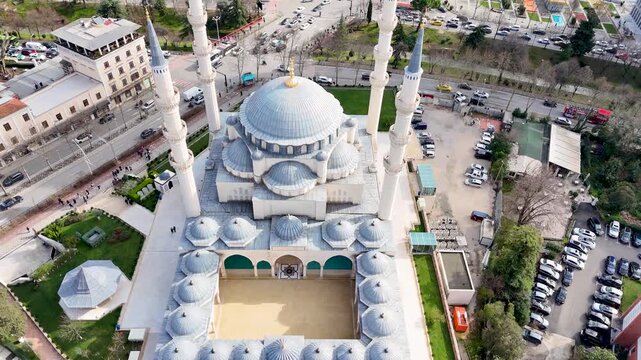 Aerial camera moves forward and tilts down toward the open inner courtyard and symmetrical domes of Namazgah Mosque in Tirana, Albania, revealing the adjacent busy street traffic