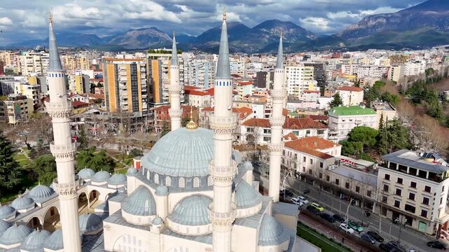 Aerial circles counterclockwise around the central dome and four minarets of Namazgah Mosque, revealing the dense city buildings and surrounding mountains of Tirana, Albania