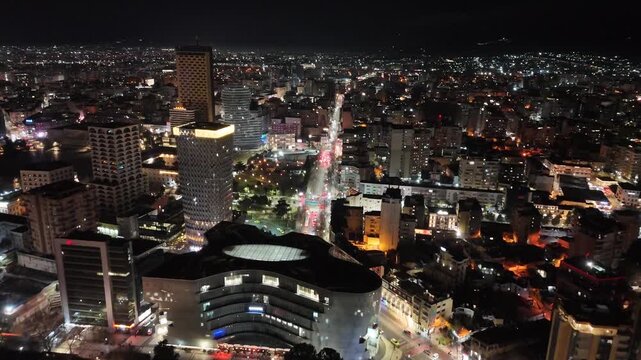 Aerial view flying forward over Skanderbeg Square park, modern curved glass buildings, and the illuminated high-rise skyline of Tirana at night