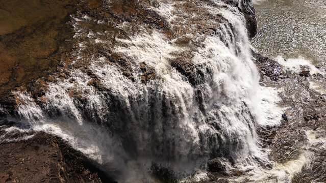Close-up aerial view of the rocky outcrop and cascading water at Sae Pong Lai Waterfall, Sekong Province, Laos. The powerful flow of water carves through the brown rocky terrain, creating dramatic pat