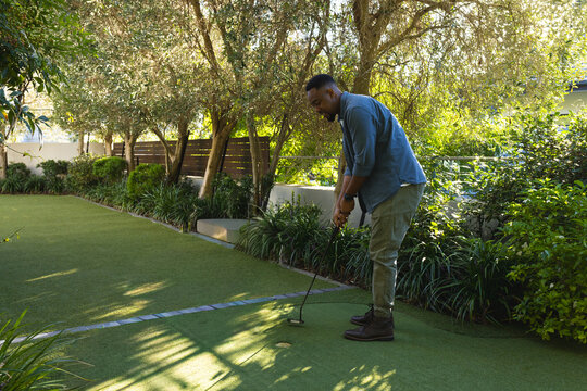 African American male wearing shirt preparing putt on yard green with putter, golf ball, copy space