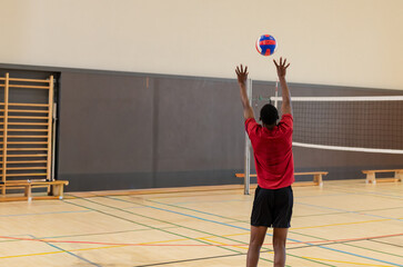 African American man jumping, reaching for red-blue-white volleyball on gym floor wearing red shirt © wavebreak3