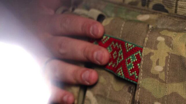 A cinematic close-up of a soldier's hand gently touching a tactical patch featuring traditional Ukrainian embroidery (vyshyvanka) on a camouflage uniform. This shot highlights the blend of modern mili