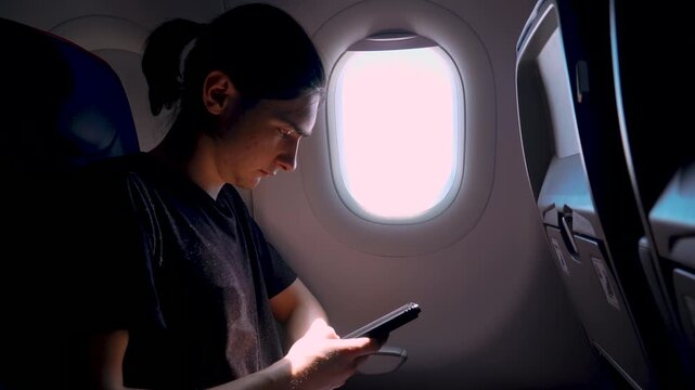Male traveler using smartphone for online banking or booking during flight, seated near aircraft window. Using mobile device during flight for digital transactions and online services. Business travel
