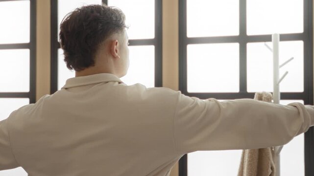 Young hispanic man with arms outstretched performing yoga t pose near large window in studio; serenity balance focus.