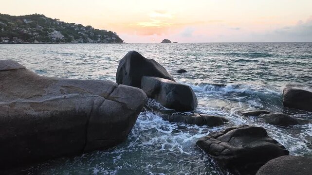 Waves crash against large coastal rocks at sunrise beneath a colorful sky