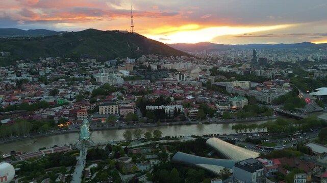 aerial view of Tbilisi, Georgia, at sunset, featuring the illuminated Bridge of Peace, the futuristic Rike Park Music Hall, and the Mtatsminda TV Tower on the mountain ridge overlooking the city