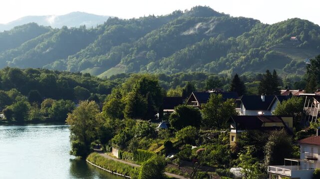 Scenic riverside neighborhood near Maribor, Slovenia with houses beside the Drava River and forested hills in warm evening light. Natural European environment during spring season.