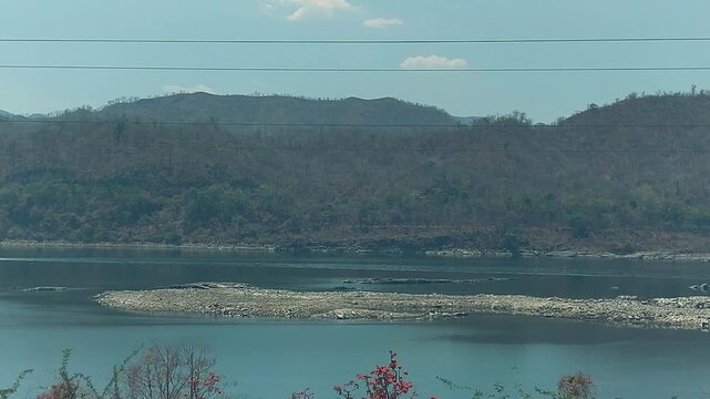 Low Narmada river exposes rocky banks amid deforested hills with flame-of-the-forest trees in foreground. Powerful for drought, water crisis, and India environmental docs.