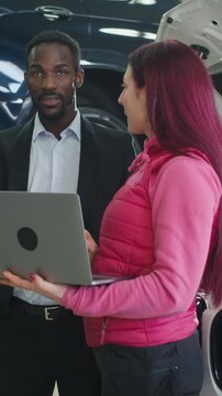 Woman mechanic showing a vehicle diagnostic result on a laptop to a male customer in a workshop with lifted cars and professional equipment.