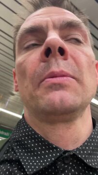 Frustrated white man speaking in subway selfie closeup under metallic ceiling lights, dotted shirt and backpack straps visible, urgent expression