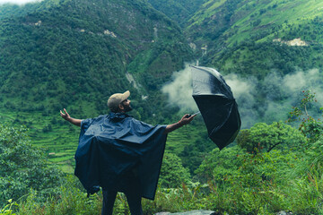 Man in a raincoat holding an umbrella stands in the lush green mountains of Ali Bedni Bugyal, Chamoli, Uttarakhand, India © 3 Travelers