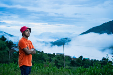 Man in an orange shirt stands looking at the cloudy mountain landscape in Ali Bedni Bugyal, Chamoli, Uttarakhand, India © 3 Travelers