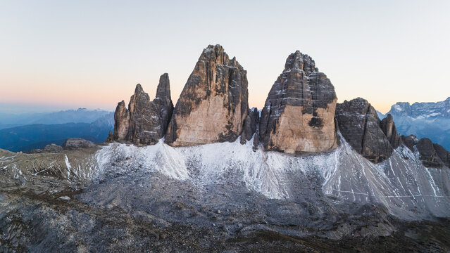 Aerial view of the iconic Tre Cime di Lavaredo peaks with a light dusting of snow on the rocky slopes under a soft twilight sky in Auronzo di Cadore, Veneto, Italy.