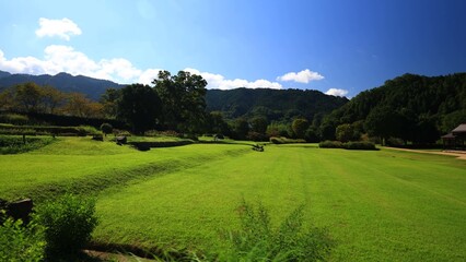 Obraz premium A spacious grassy field stretches toward forested hills with two open wooden pavilions standing in sunlight beside a winding dirt path creating a calm natural setting of balance and rural simplicity 