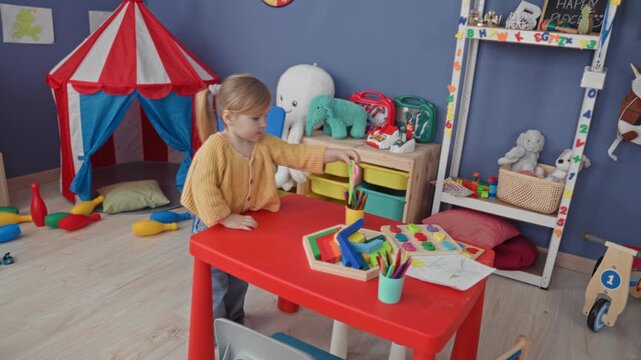 Girl holds scissors by red table amid wooden toys and small tent in a playroom inside building; curiosity.