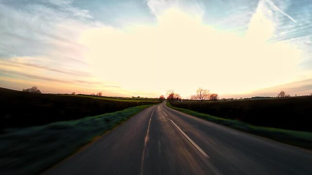 Traveling along winding countryside road across farmland at sunset, rural driving journey first person view. Curved asphalt lane moving through open fields under warm sky light, evening road trip
