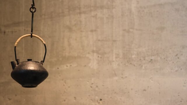 Black cast iron kettle hanging by metal handle on a hook in warm light. Rustic historical style object used for fire heating and traditional cooking against a concrete wall background