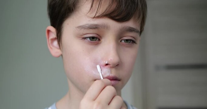 Child applying cream on face. Young boy uses cotton swab for skincare routine. Morning routine emphasizes self-care and hygiene. Calm atmosphere creates focus on skincare.