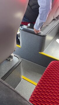 Caucasian commuter secures suitcase on bus, hand holding handle near stepwell and armrest, red patterned seat and yellow safety strips visible, steady