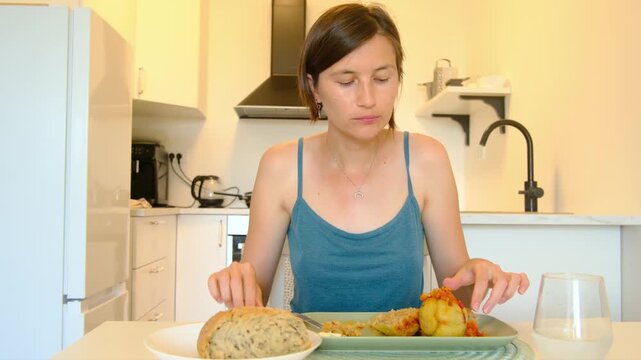 Woman sits at kitchen table eating stuffed paprika, enjoying homemade Balkan cuisine, turning meal into cultural moment shared in quiet domestic space.