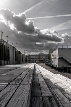 Monochrome promenade runs beneath clouds through urban architecture, with building edges and exterior daylight perspective stretched across long decking