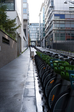 Rows of bicycles line a wet sidewalk for urban transportation, beside architecture and building fronts in exterior daylight on a narrow street commute