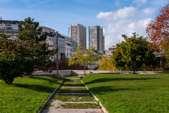 Urban park pathway crosses green grass toward architecture and distant towers, with building edges framing an exterior daylight skyline