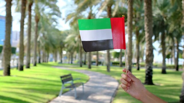 Female hand holding small UAE flag against Corniche Walking Trail in Abu Dhabi. Blend of nature, patriotism, and serenity, with lush greenery , views symbolizing harmony and progress.