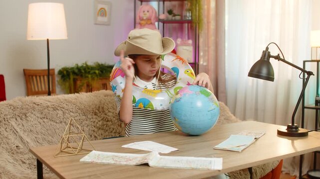 Happy child girl sitting at home table wears a travel hat and inflatable ring while looking at a globe, imagining a beach vacation. School kid enjoys dreaming about travel planning a seaside holiday