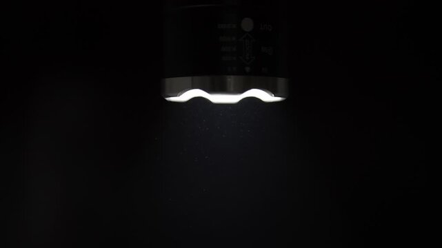 A bright LED flashlight beam shines downwards against a solid black background, revealing microscopic dust particles and motes swirling and floating in the air. This visual captures the