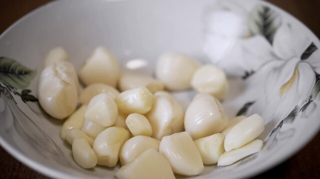 Freshly peeled garlic cloves drop slowly into a white ceramic bowl decorated with flowers. This cooking preparation concept highlights essential ingredients for flavorful seasoning.