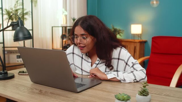 Indian woman at home types on laptop then smiles after reading reply win notification from online game. Arabian girl at table raises fist celebrating great success joyful reaction to winning message.