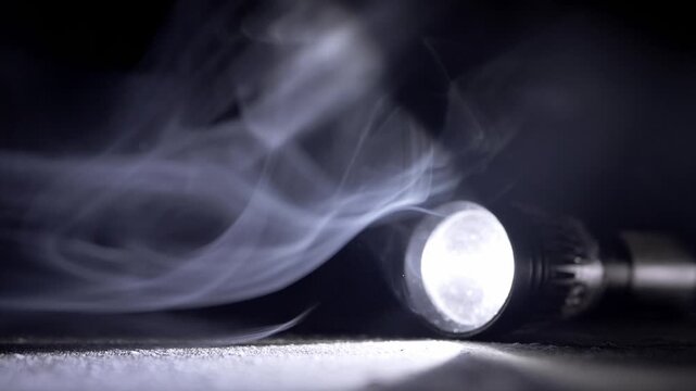 A bright tactical flashlight lies on a dark surface projecting a beam through thick swirling white smoke. Concept of mystery, emergency rescue, and finding direction in deep darkness.