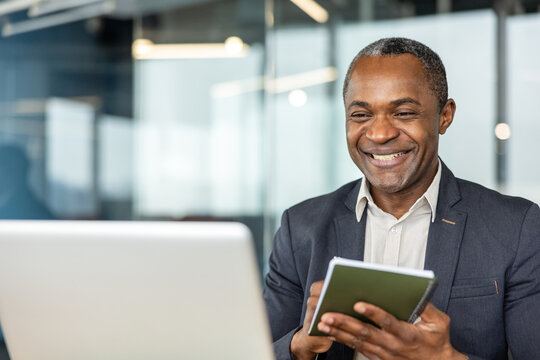Mature professional man in a modern office smiling during a virtual meeting on his laptop while taking notes in a notebook, demonstrating confident remote collaboration and productivity