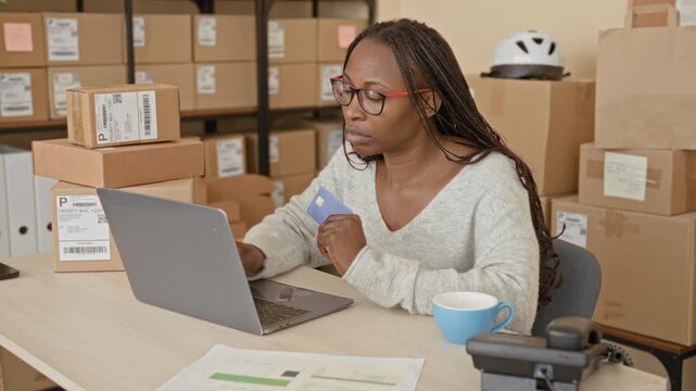 Woman holding creditcard and typing on laptop in a shipping area of a small business building with stacked parcels and mailing labels; determined small business owner.